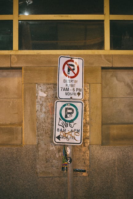 Close-up of a vertical parking restriction sign mounted on a metal pole attached to a stone wall, with a large window with multiple panes partially visible above. The top sign indicates no parking except by permit from 7 am to 6 pm Monday to Friday, with a red circle and slash over a black 'P' symbol on a white background; the lower sign specifies restricted parking hours from 8 am to 6 pm Monday to Friday, featuring a blue circle with a white 'P' and a hand symbol, indicating permit or resident parking. The signpost appears to be located in an urban setting, possibly outside a residential or commercial building, with a concrete pavement area at the base. The image is relevant to house removals and relocation services provided by Man with Van Belvedere, as it depicts parking regulations that may impact loading and unloading during home moves or furniture transport.