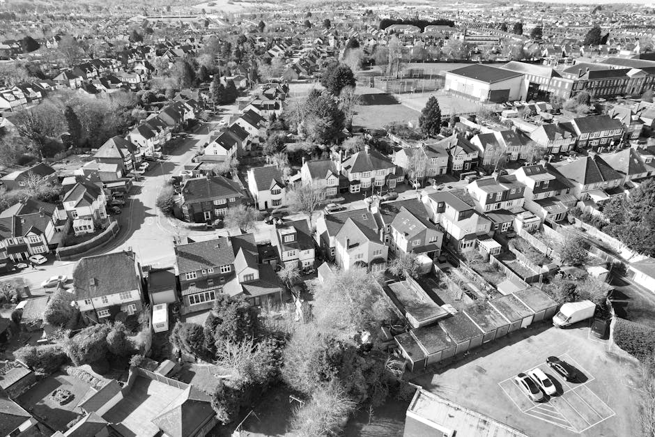 An aerial black and white photograph showing a residential neighbourhood with a mix of detached and semi-detached houses, many with pitched roofs and small gardens. The houses are closely spaced along winding streets, with trees and shrubbery dispersed throughout. In the lower right corner, there is a paved parking area with several parked cars and a white van, likely used for home relocation or furniture transport. Adjacent to the parking lot, a fenced yard contains flat-roofed structures, possibly sheds or garages, with visible pathways and some gardening elements. In the background, there is an open sports field or schoolyard, bordered by trees and additional housing. The image captures the scene during daylight, with natural lighting highlighting the various structures and greenery. The photograph contextualizes the process of packing and moving, with [COMPANY_NAME]'s removals service potentially involved in transporting furniture and household items across this suburban setting in Belvedere, Bexley.