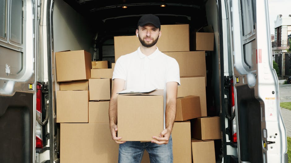 A white moving van from Man with Van Belvedere is parked on the street in front of a residential building with a brick and white facade. The van has the company logo and the words 'MOVING COMPANY LOCA