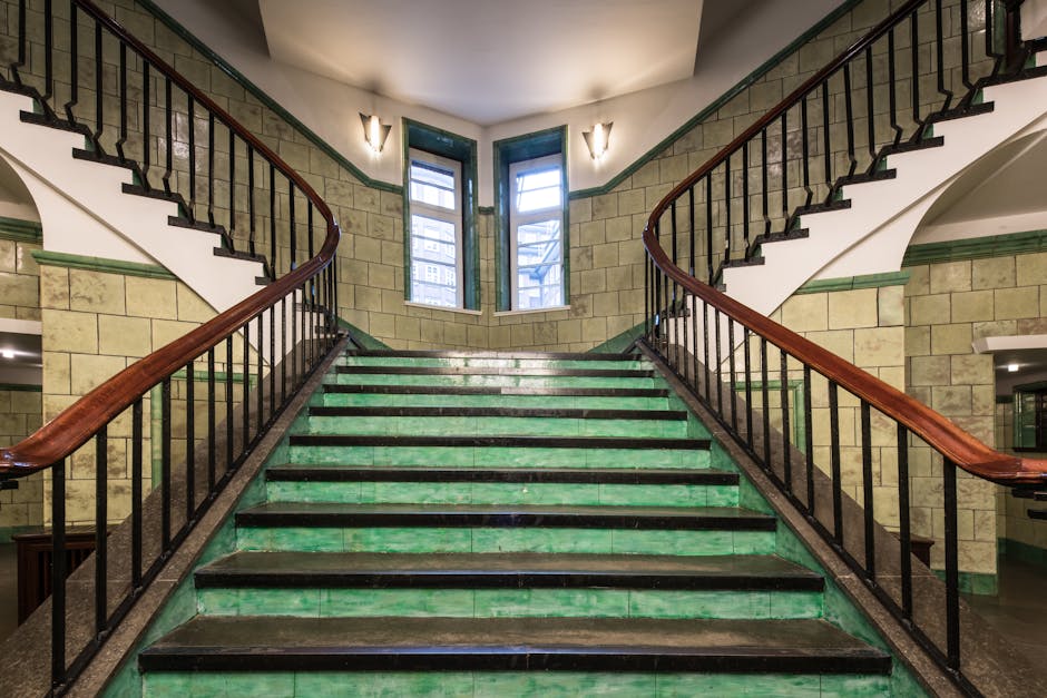A wide staircase inside a building with green-tiled steps and black anti-slip strips along the edges, leading up to a small landing with three windows at the top allowing natural light to illuminate the area. The staircase has dark wooden handrails on both sides supported by black metal balustrades. The surrounding walls are constructed with beige stone bricks, with green trim along the top edge. The interior setting appears to be part of a residential or commercial property, featuring classic architectural details. This environment represents a location where home relocation or furniture transport logistics might take place, as seen in the context of house removals and the associated staircase access. Man with Van Belvedere may use this staircase during the process of loading furniture or boxes for moving services, supported by the natural lighting and spacious design suitable for moving activities.