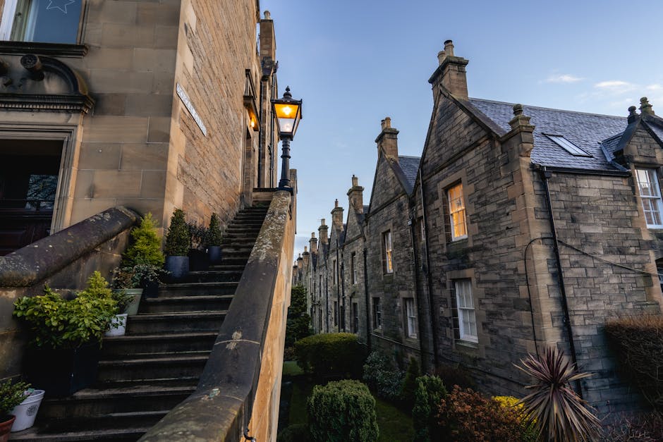 Exterior view of a residential property with stone walls and pitched slate roofs, showing a staircase with a stone balustrade on the left side leading from the street level to an entrance illuminated by a vintage-style lantern. Several potted plants are placed along the staircase, and the surrounding garden has neatly trimmed bushes and ornamental plants. Adjacent to the building, a row of similar stone-built houses extends into the background, with multiple chimney stacks visible on the rooftops. The scene is captured during early evening with a clear sky, and a van may be visible nearby for home relocation or furniture transport, reflecting the context of house removals and moving services by Man with Van Belvedere.