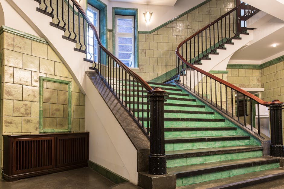 A wide staircase inside a building with green-tiled steps and black anti-slip strips along the edges, leading up to a small landing with three windows at the top allowing natural light to illuminate the area. The staircase has dark wooden handrails on both sides supported by black metal balustrades. The surrounding walls are constructed with beige stone bricks, with green trim along the top edge. The interior setting appears to be part of a residential or commercial property, featuring classic architectural details. This environment represents a location where home relocation or furniture transport logistics might take place, as seen in the context of house removals and the associated staircase access. Man with Van Belvedere may use this staircase during the process of loading furniture or boxes for moving services, supported by the natural lighting and spacious design suitable for moving activities.
