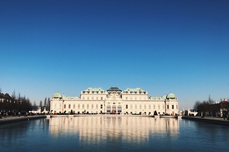 A large, historic white building with multiple green-domed roofs and ornate architectural details, situated behind a reflective water feature that mirrors the structure. The scene is outdoors with a clear blue sky overhead and a paved area in front of the building. Few people are visible around the perimeter, likely taking part in a home relocation or furniture transport process. The building appears to be part of a park or civic space, with trees lining the background. Inside the water feature, there may be some floating or submerged objects, while the area in front of the building is used for loading or unloading furniture, boxes, or appliances during a house move. Man with Van Belvedere occasionally assists with relocating household items and furniture, supporting efficient packing and moving operations, especially in urban or historic settings such as this.