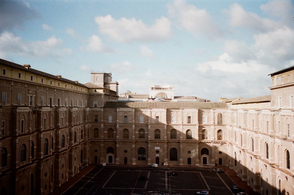 An outdoor view of a large, historic, multi-storey building with a central courtyard, captured during daytime with partly cloudy skies. The building features numerous evenly spaced rectangular windows with decorative arches and stone frames, set against beige or light brown stonework. The rooftop shows a mix of flat and slightly pitched sections, with some structures or equipment visible on top. In the foreground, the courtyard is paved with arranged tiles or stones, and there are a few parked cars along the edges. The scene suggests an urban environment potentially suitable for home relocation or moving services, with the building's spacious layout implying interior furniture and belongings could be transported via nearby loading areas or adjacent streets, supported by companies like Man with Van Belvedere specializing in removals and transport logistics.