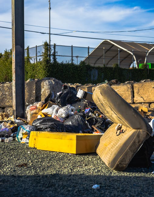 An outdoor scene showing a pile of bulky waste and household rubbish, including black plastic garbage bags, cardboard boxes, and a detached car seat cushion, placed on a gravel surface near a stone wall. A yellow plastic container is also visible among the waste. In the background, there is a metal fence, some greenery, and a large, curved industrial-style building under a blue sky with scattered clouds. The waste is situated adjacent to a paved walkway, likely part of a home relocation or clearance process, with no vehicles or moving equipment immediately visible. The setting indicates a disposal area related to a house move, with Man with Van Belvedere potentially assisting with clearance or waste removal services involved in the process of packing, loading, and transporting items during a house removal or furniture transport operation.