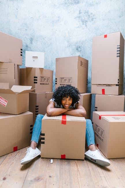 A young woman with curly black hair is sitting on the wooden floor inside a room, surrounded by numerous cardboard boxes of various sizes, some sealed with red and black packing tape and others partly