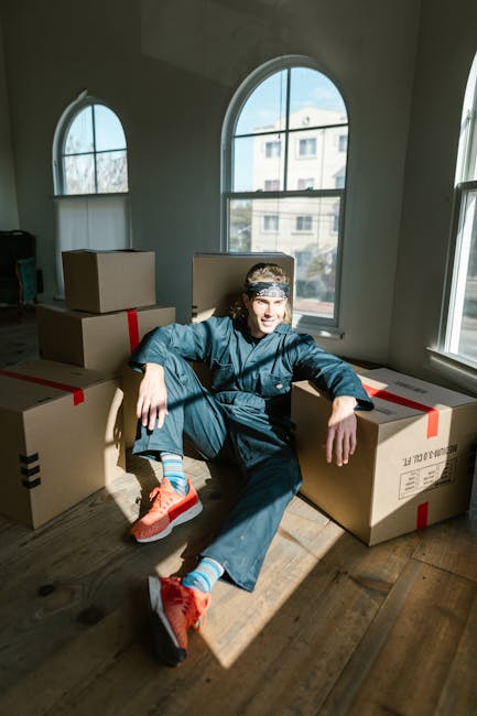 A young woman with curly black hair is sitting on the wooden floor inside a room, surrounded by numerous cardboard boxes of various sizes, some sealed with red and black packing tape and others partly