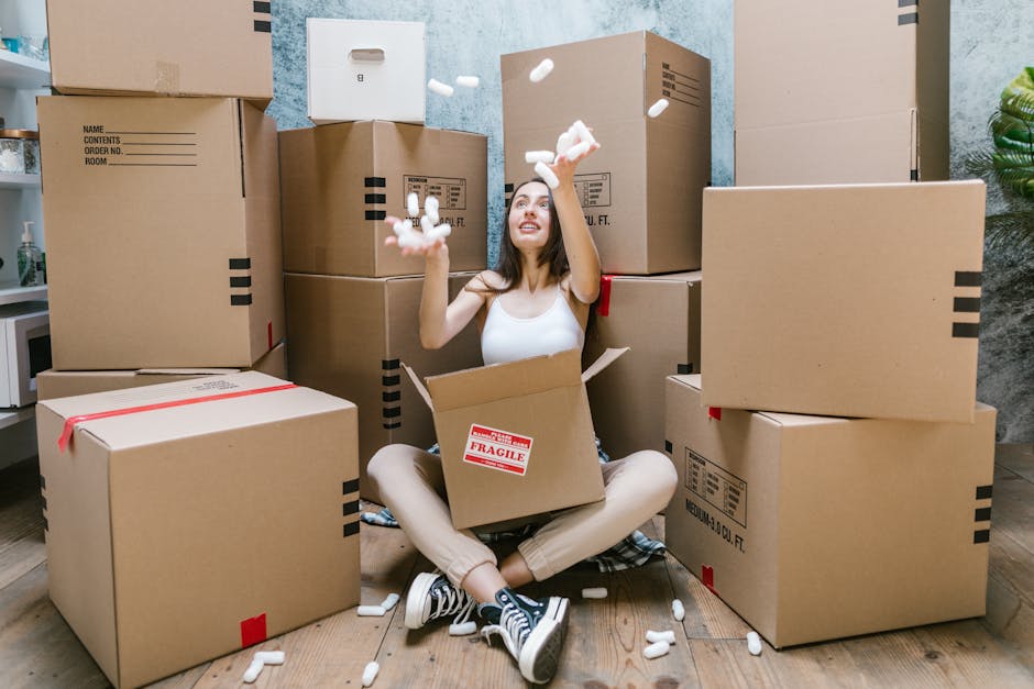 A young woman with long brown hair, dressed in a white tank top, beige pants, and black sneakers, is sitting on a wooden floor amidst numerous unpacked cardboard boxes of varying sizes, some sealed with red tape and others open, revealing packing materials such as white foam peanuts. She is smiling and tossing foam peanuts into the air, appearing to be in the process of packing or unpacking during a home relocation. The scene is set indoors, with a blue textured wall behind her, a small white shelf with a potted plant partially visible on the right, and natural lighting illuminating the space. Several cardboard boxes are stacked around her, indicating an active packing or moving process facilitated by Man with Van Belvedere, a local removals service supporting furniture transport and home moving logistics.