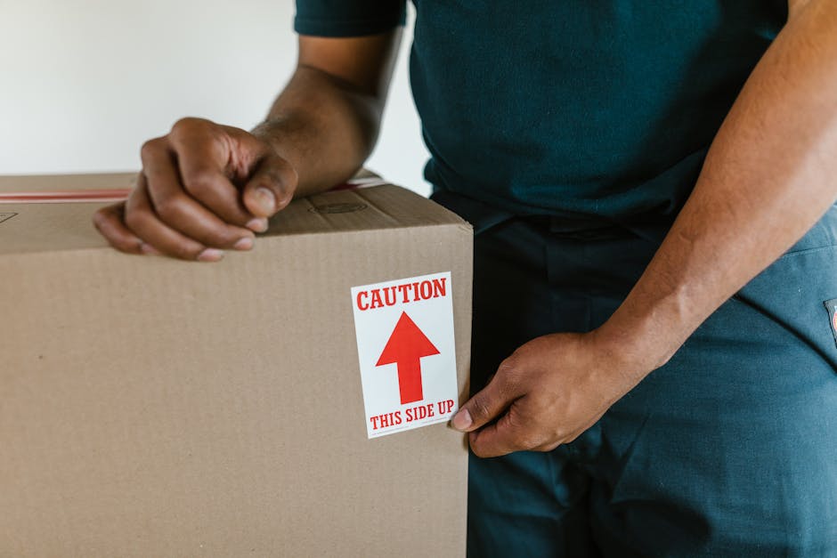 Close-up of a person wearing a dark green T-shirt and blue trousers during home relocation, attaching a red and white caution label that reads 'CAUTION THIS SIDE UP' with an upward arrow onto a large cardboard moving box. The box is positioned inside a property, likely in a doorway or near an entry point, with the person's hands securing the label, indicating careful handling during packing and moving processes. In the background, part of a light-colored wall and the edge of a door frame are visible, suggesting indoor environment. The scene reflects the packaging stage of furniture transport and packing for a professional removal service, with emphasis on safety and proper handling during house removals. Occasional references to [COMPANY_NAME] and their expertise in providing efficient house removals and loading services are implied through context, aligning with the page about same-day emergency moves in Belvedere.