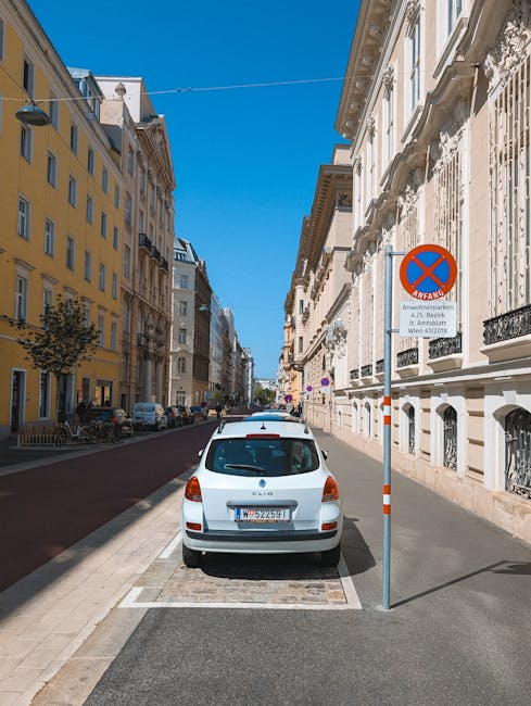 A white hatchback car parked in a designated parking space on a narrow city street with historic, multi-story buildings on both sides. The vehicle faces away from the viewer, with visible rear lights and a European license plate. To the right of the car, a blue and red circular parking restriction sign is mounted on a metal pole, indicating restrictions for parking enforcement. The sign includes additional information about parking permits or restrictions, with a background of blue sky and bright sunlight illuminating the scene. The street surface is a mix of asphalt and paving stones, with a curb separating the parking area from the main road, and other parked cars can be seen further down the street. This setting reflects an urban environment suitable for facilitating house removals or furniture transport, as managed by companies like Man with Van Belvedere, who support packing and moving logistics in residential areas.
