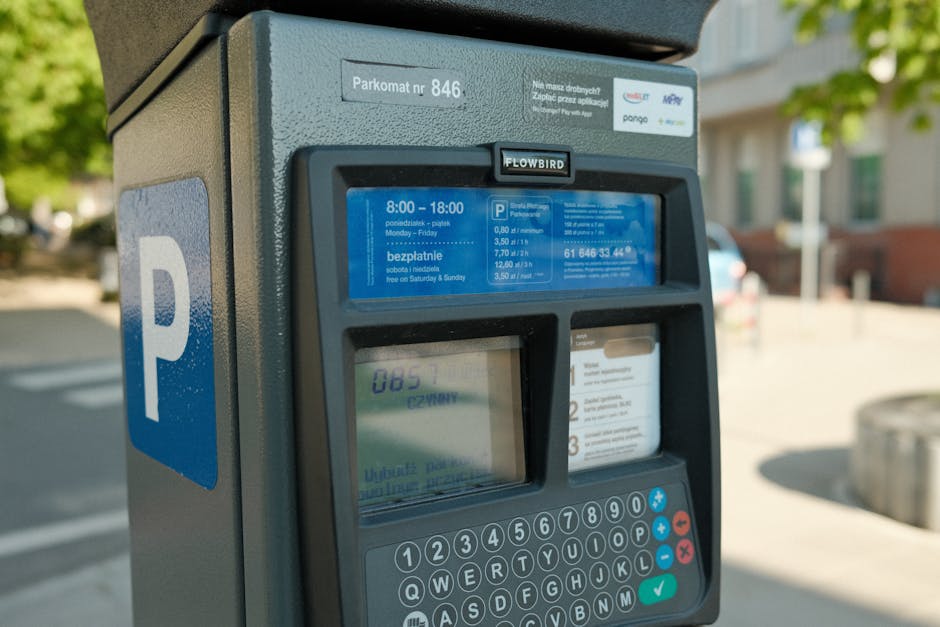 A close-up image of a parking meter situated on a sidewalk in a residential area, with a blue parking sign displaying a white 'P' in the background. The parking meter features a digital screen showing parking details and costs, a keypad with numeric and function buttons, and a small card reader slot. The surrounding environment includes blurred trees, a building, and a parked car, highlighting an outdoor setting during daylight. This scene exemplifies the parking management process often encountered during home relocation or furniture transport when managing parking permits, aligning with services offered by Man with Van Belvedere, particularly concerning house removals and moving logistics.