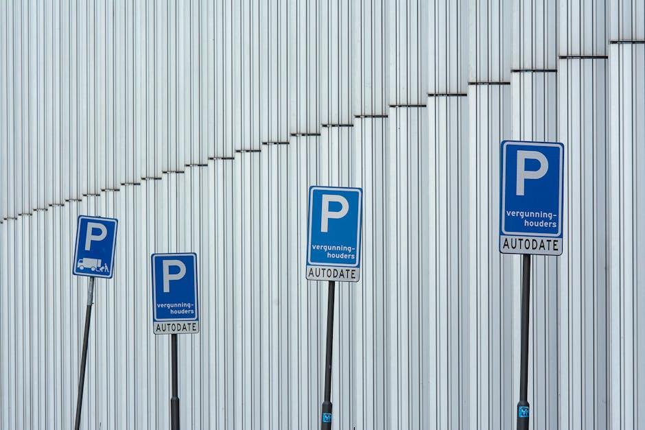 A white hatchback car parked in a designated parking space on a narrow city street with historic, multi-story buildings on both sides. The vehicle faces away from the viewer, with visible rear lights and a European license plate. To the right of the car, a blue and red circular parking restriction sign is mounted on a metal pole, indicating restrictions for parking enforcement. The sign includes additional information about parking permits or restrictions, with a background of blue sky and bright sunlight illuminating the scene. The street surface is a mix of asphalt and paving stones, with a curb separating the parking area from the main road, and other parked cars can be seen further down the street. This setting reflects an urban environment suitable for facilitating house removals or furniture transport, as managed by companies like Man with Van Belvedere, who support packing and moving logistics in residential areas.